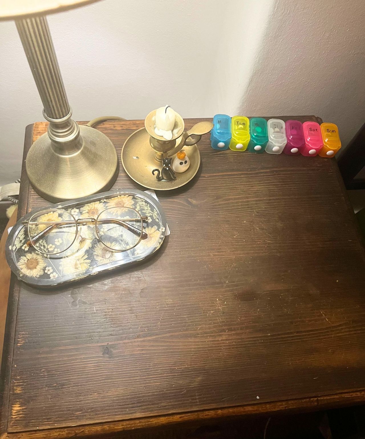 The top of a dark wooden nightstand with a lamp, a pair of glasses, a pill box, and a candle holder