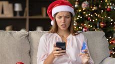 Stressed woman in Santa hat sitting in front of a Christmas tree making purchases on her phone while holding a credit card