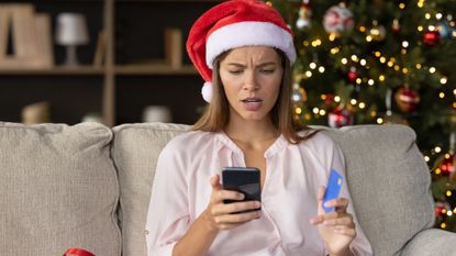 Stressed woman in Santa hat sitting in front of a Christmas tree making purchases on her phone while holding a credit card