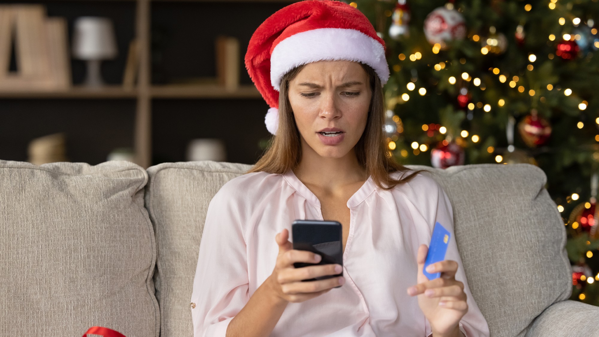 Stressed woman in Santa hat sitting in front of a Christmas tree making purchases on her phone while holding a credit card