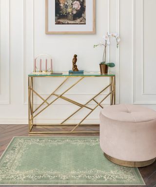 An entryway with a pistachio green rug, a pale pink storage ottoman, a gold and glass table with candles and an orchid on top, and white wall paneling