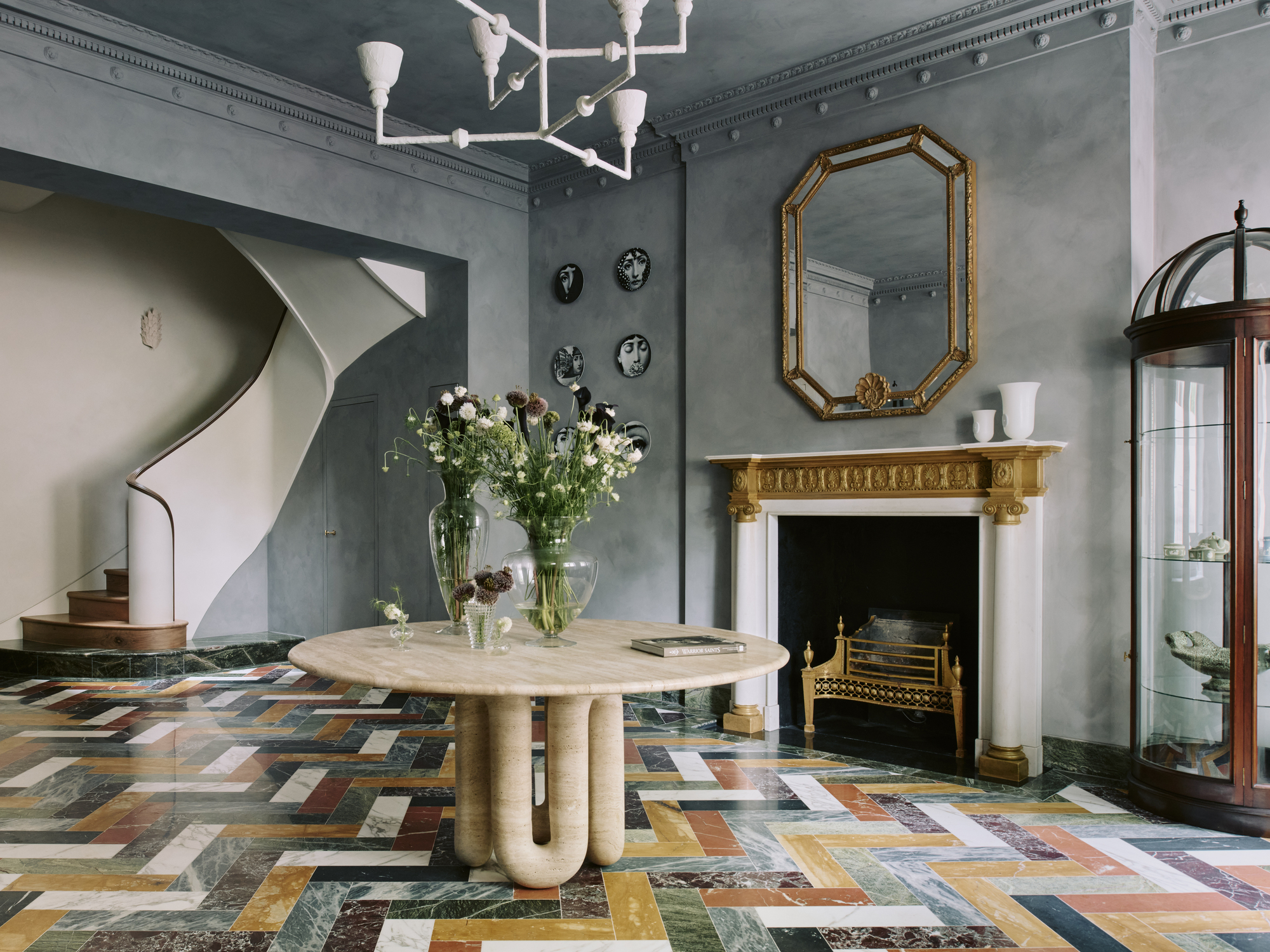 Image of a grand entrance hall that has limewashed, powder blue walls, and colorful tile floors. There is a white and gold fireplace and a mirror hanging over it.