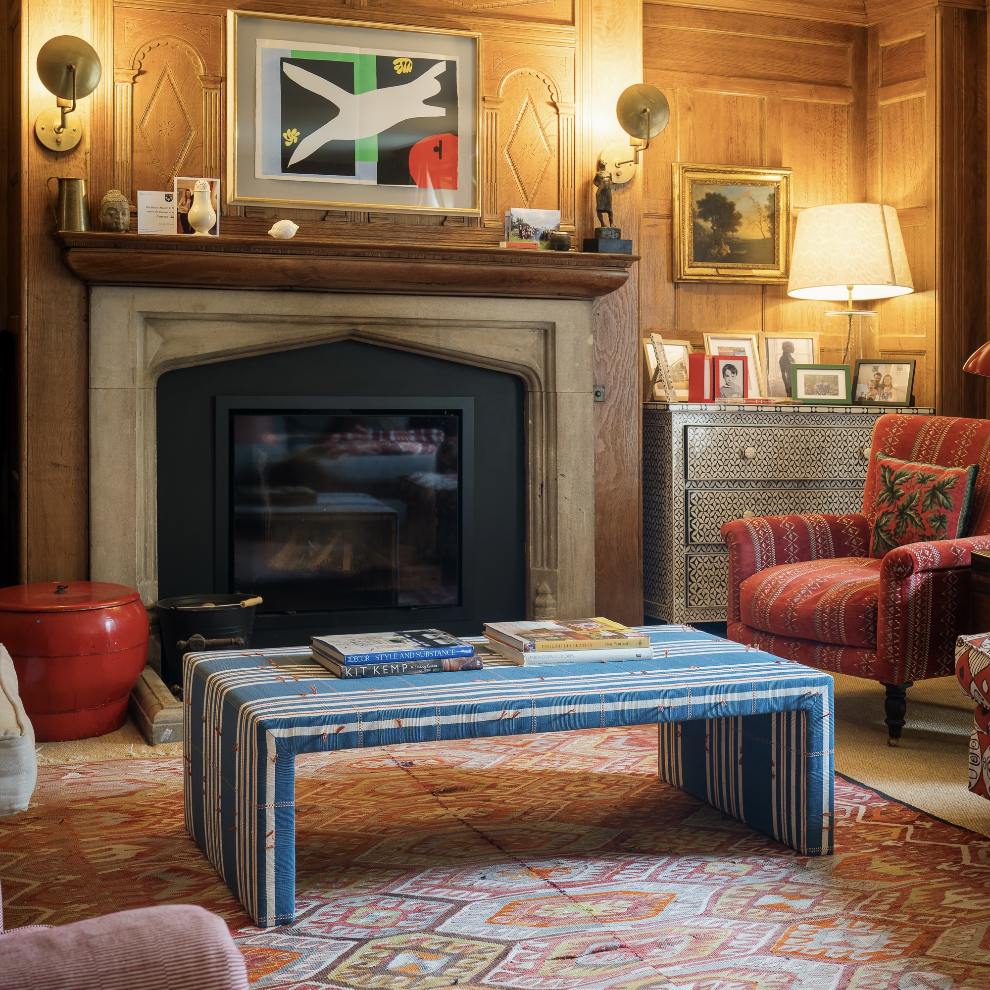 Wood panelled living room with large stone fireplace, patterned rug on floor and upholstered patterned ottoman as coffee table