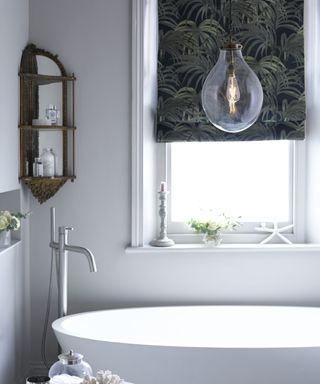 A large white bath next to a window with a dark blind and underneath a large pendant light
