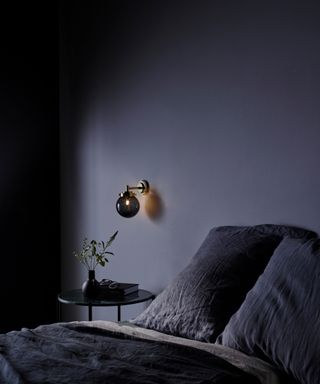 Dark and moody bedroom detail, with glass orb wall light above bedside table, and lived-in crumpled bedlinen in deep indigo colorway.