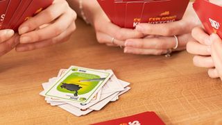 A group of people hold Exploding Kittens cards, with a pile between them on a wooden table