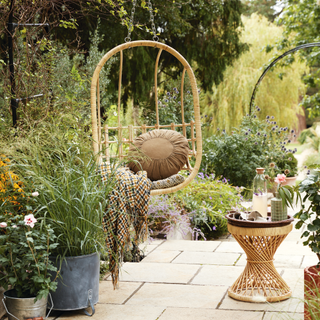 Paved patio and path with flower beds and pots of plants, hanging chair with cushions and throw.