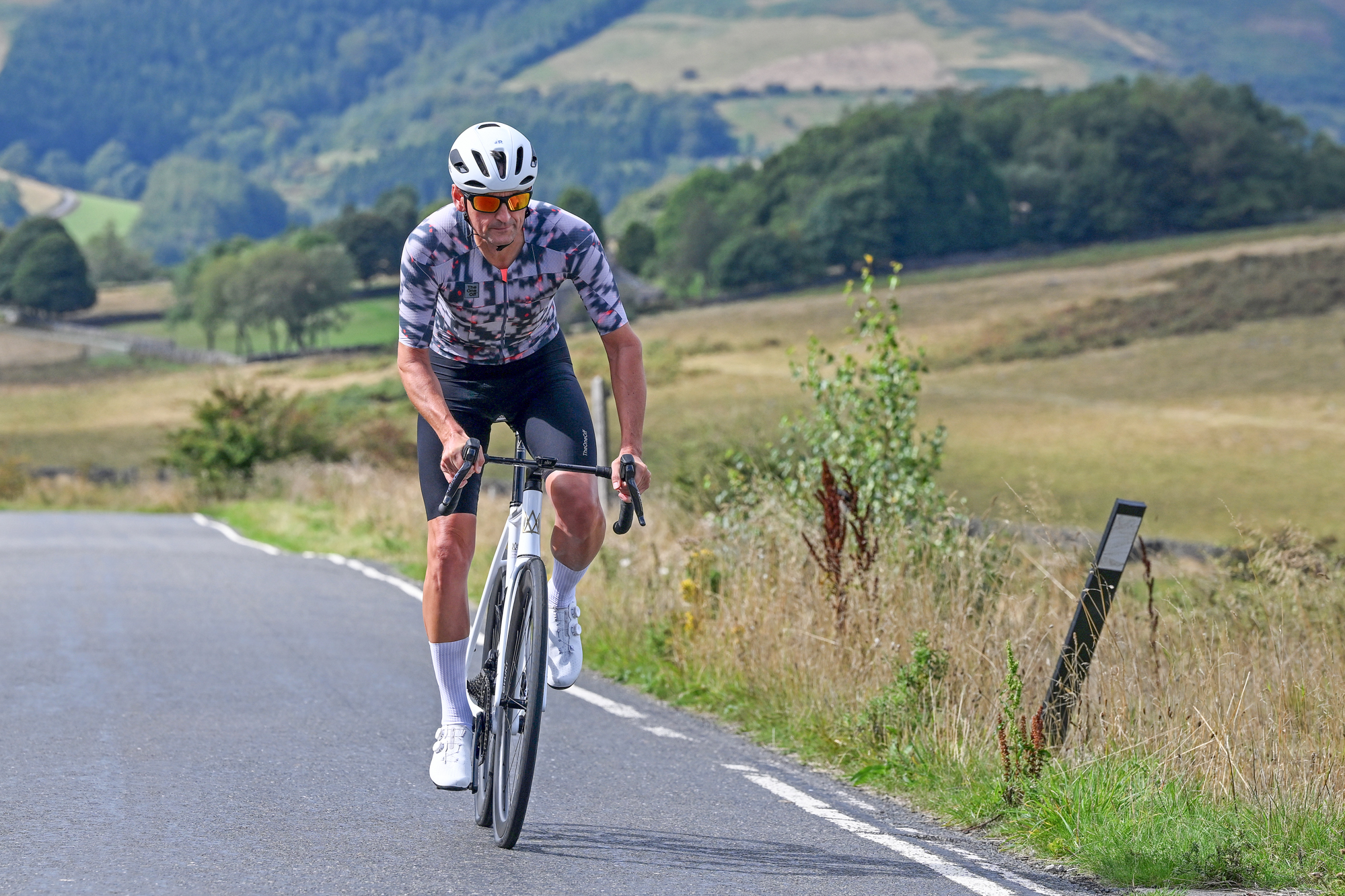 a white road bike being ridden out of the saddle towards the camera by a man wearing white shoes, white helmet, black shorts and a patterned grey jersey with green countryside hills in the background