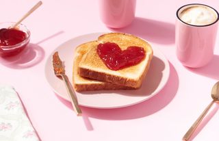 A Valentine's Day breakfast set up with toast covered in a heart-shaped jam on a pink background.