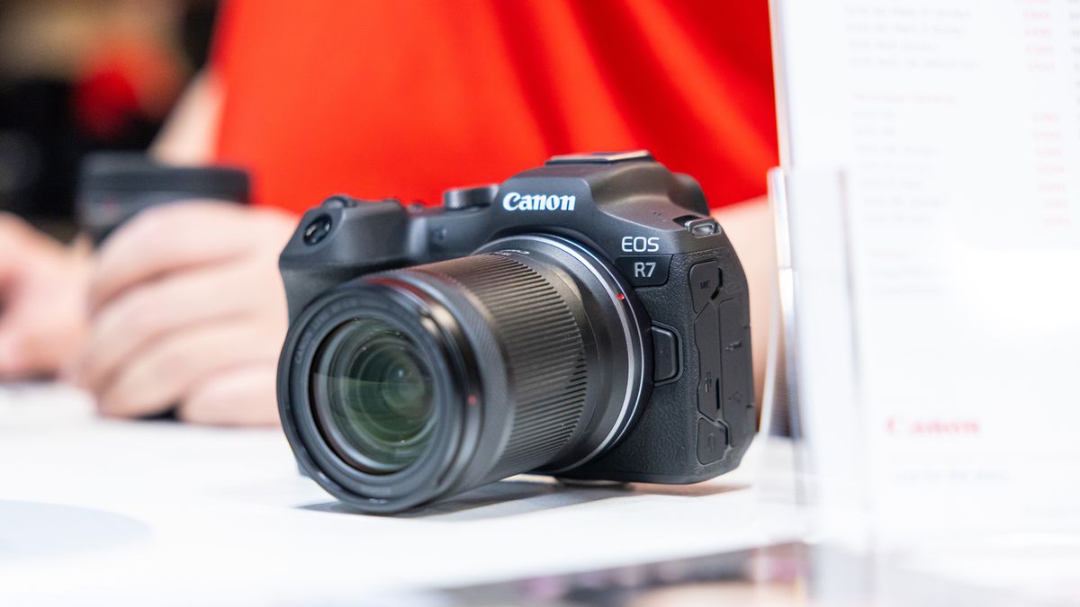 A Canon EOS R7 camera, on a white surface, in front of a Canon expert wearing a red shirt