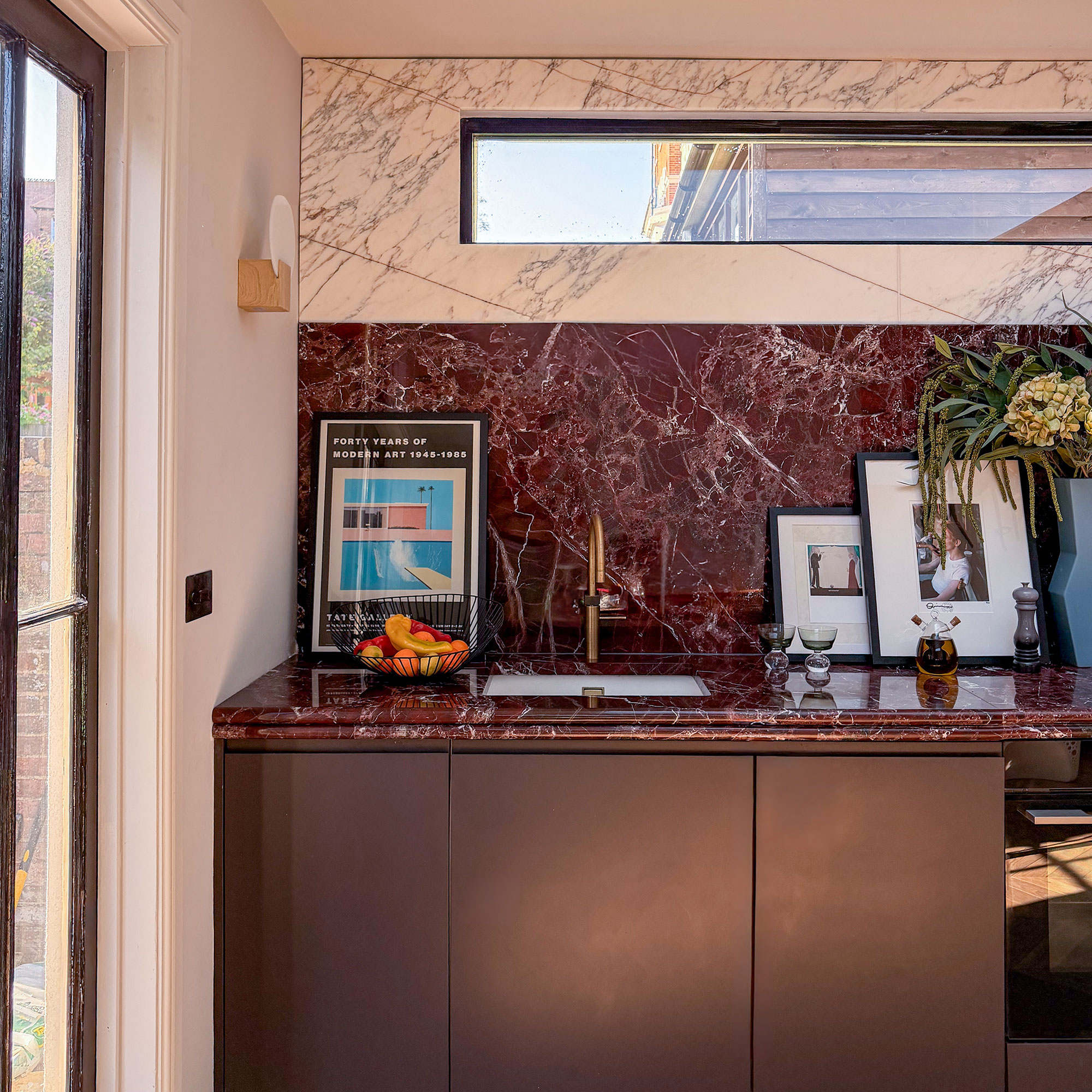 Kitchen with red marble worktops and splashbacks, topped with white marble. Dark red cabinetry. High narrow window. Photographs and vases on countertop