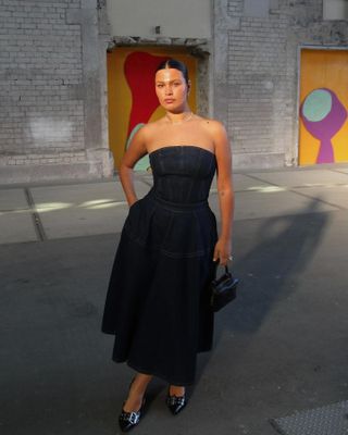 Mid-size model Bella Cohen poses at an event wearing a strapless dark denim dress, a mini black bag, and buckled pointed-toe pumps.