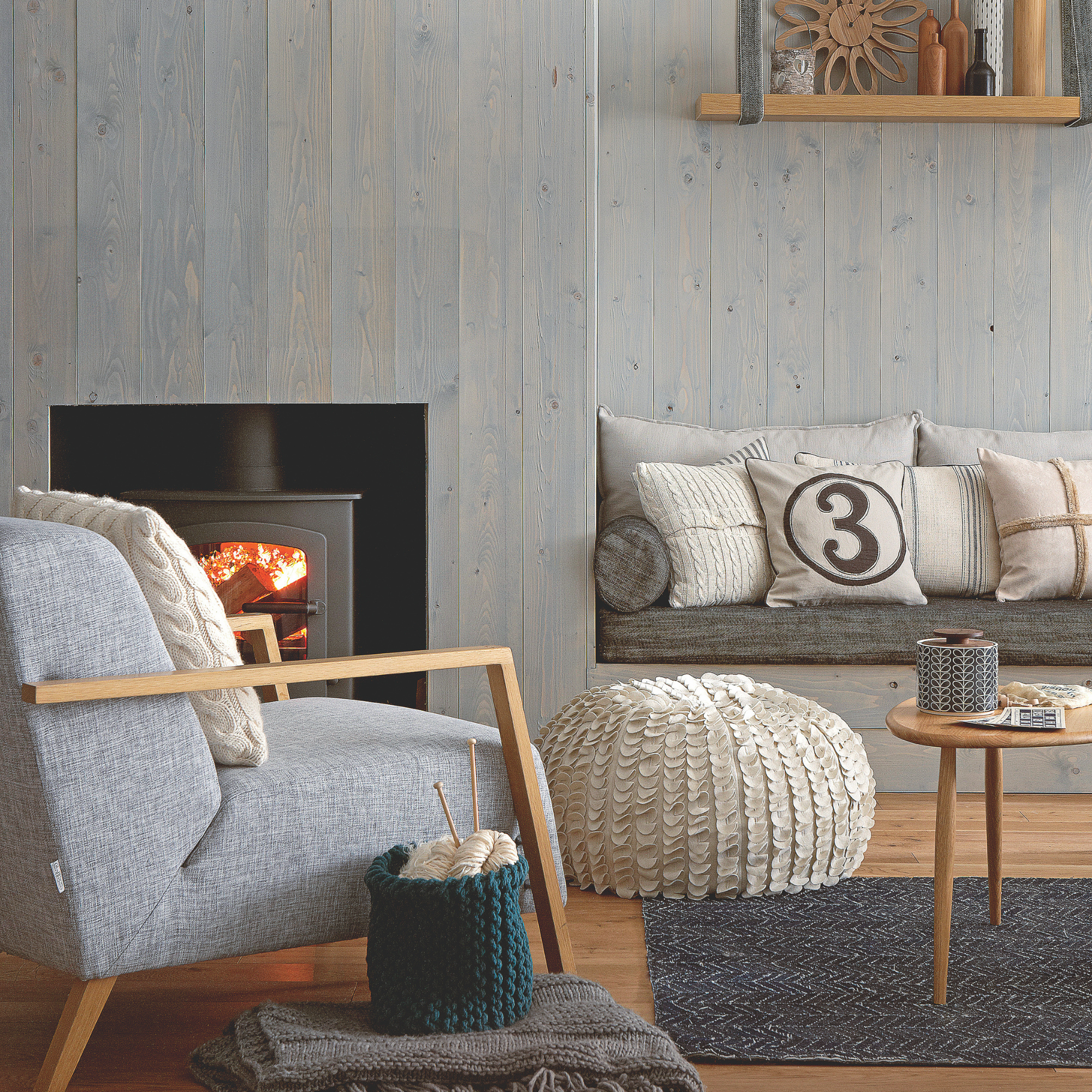 Pale blue grey pine plank wooden panelled walls, Shoreditch E.1 sign post, lit wood burner, stove wooden framed grey upholstered armchair, knitting kit, cream petal effect beanbag, wooden bench with cushioned seating, number 3 cushion, wooden shelf hung with material