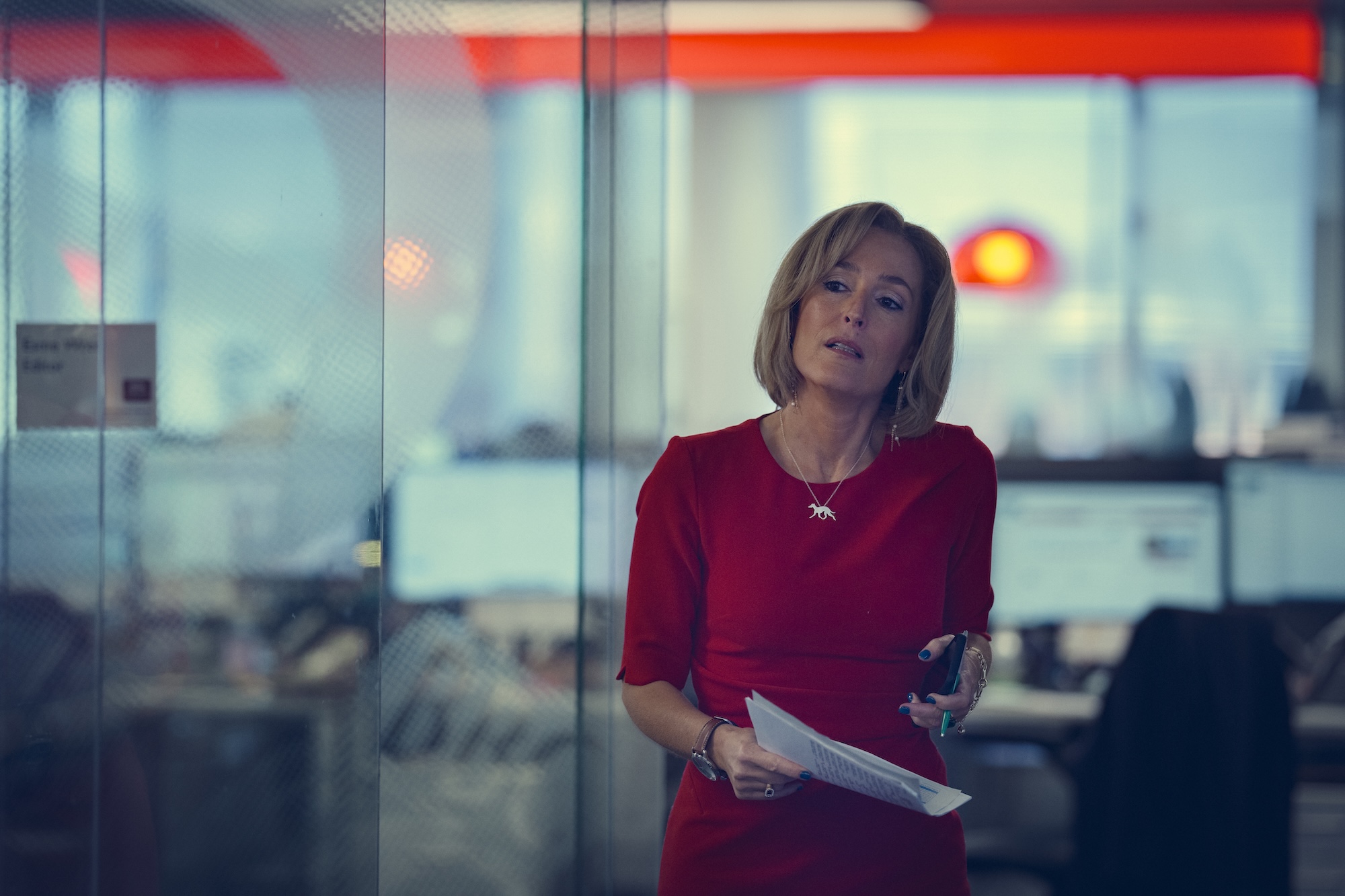 a woman in a red dress (gillian anderson as emily maitlis) holds papers and a phone while standing in a glass-walled office, in the netflix movie 'scoop'