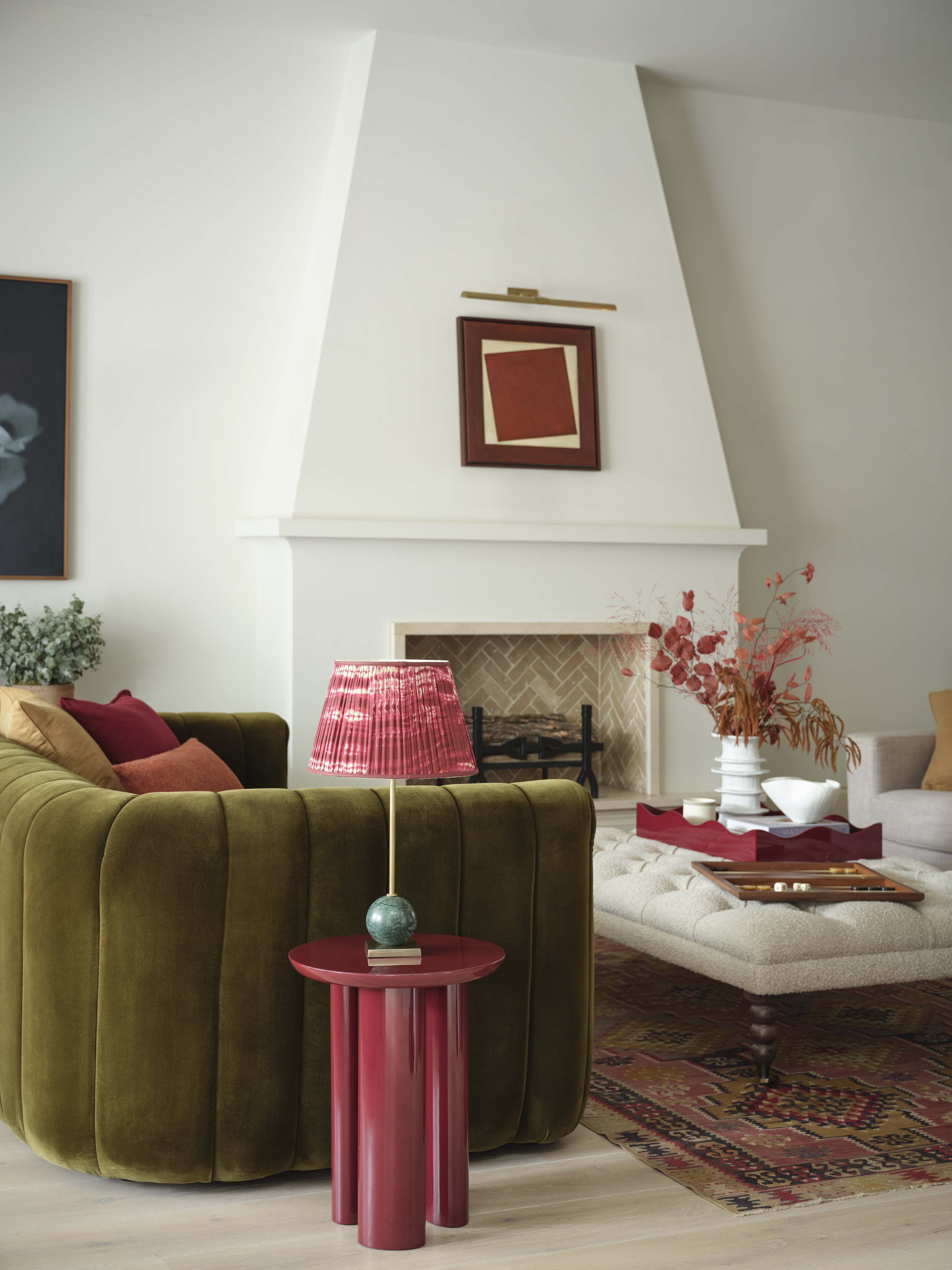 a living room with a green velvet sofa and large fireplace with artwork on, a table lamp is on a table in the foreground