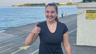 Emery Little poses for a photo after a run near a large body of water. Her hair is tied back and she is wearing a sports t-shirt and she gives a thumbs-up to the camera.