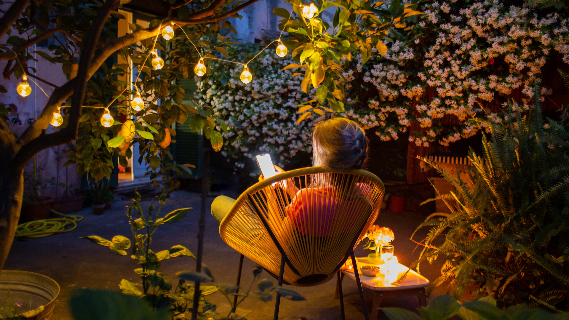 A woman reading her phone in a chair in her garden, at dusk