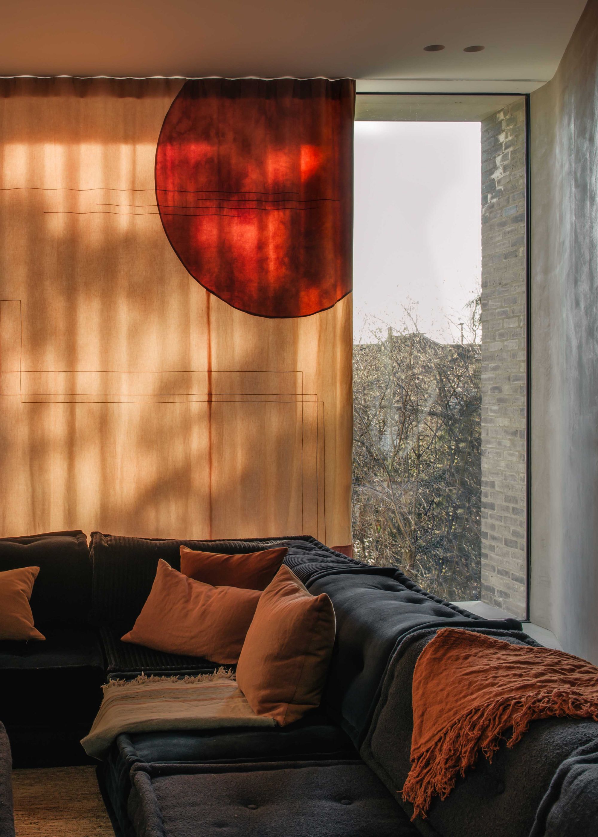 The corner of a modern living room with a floor-to-ceiling window with a peach colored curtain with an abstract red print. A large dark blue corner sofa with warm rust-colored cushions and throws.