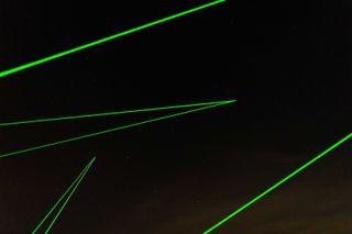 Night image, dark cloudy sky, four sets of green laser beams projected across the sky from ground level, of the space waste lab tracking system