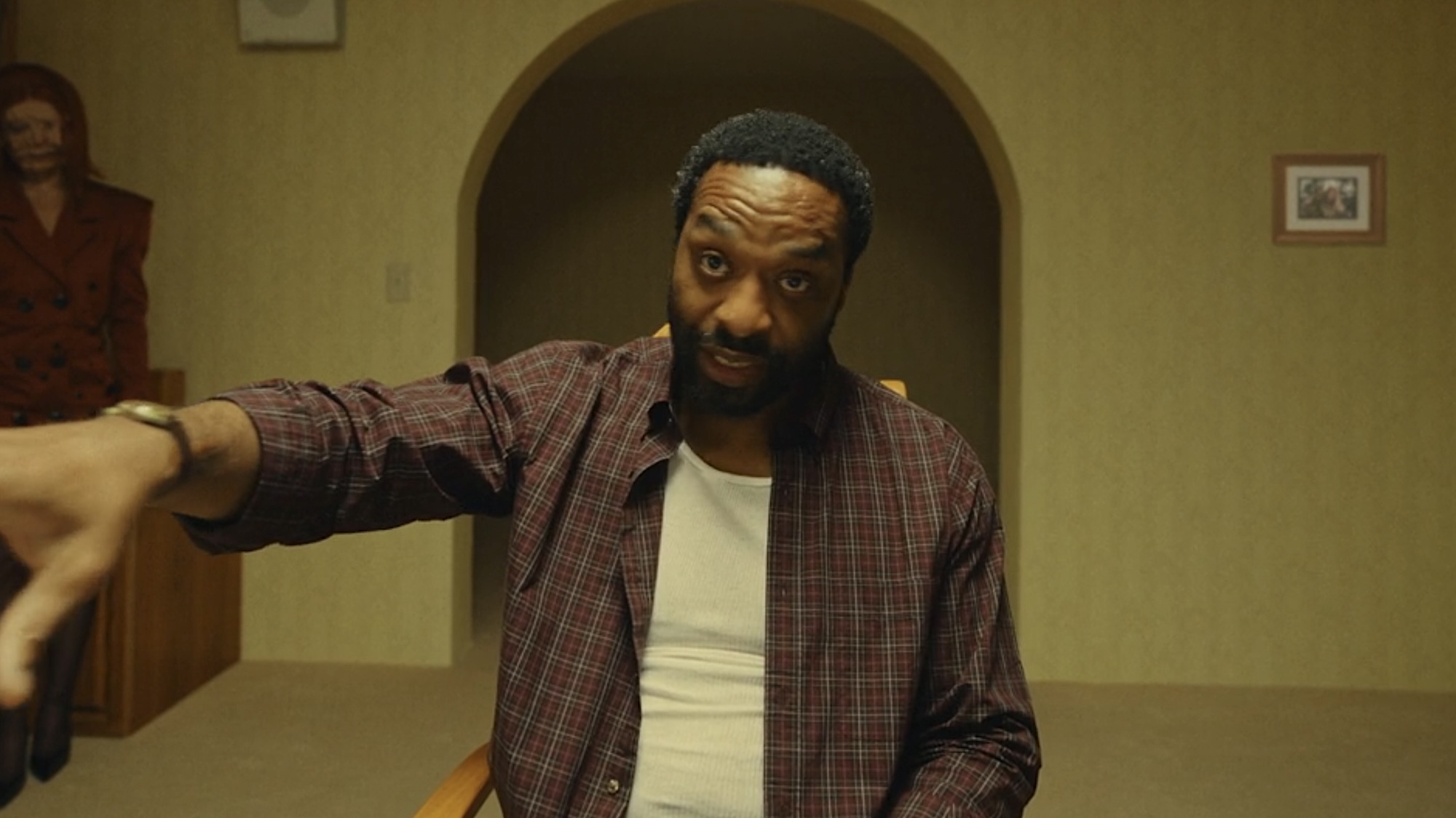 Chiwetel Ejiofor sitting in checkered overshirt and white T-shirt, holding hand out to point to male Still-Life, as redheaded woman still-life stands behind him and to the side. 