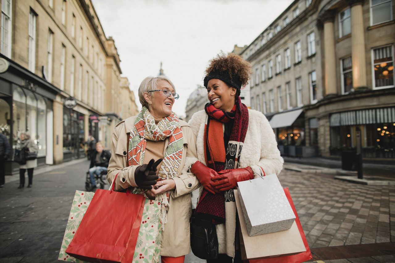 Two older women carrying bright shopping bags are walking down a commercial street and talking together.