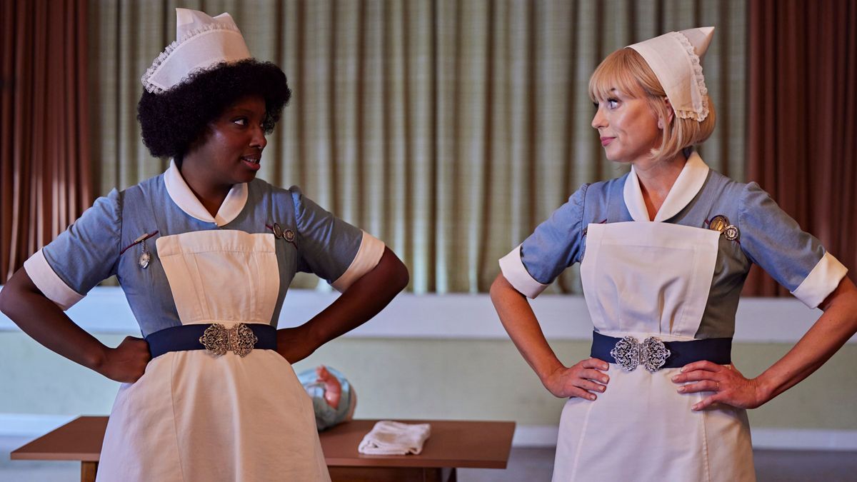 Joyce and Trixie side by side in their midwife uniforms as they look at one another with their hands on their hips in Call the Midwife season 14