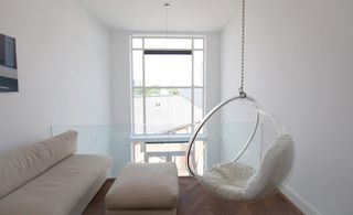 Room featuring white wall, dark wood flooring and glass panel. Long cream couch against the white wall with cream footrest and an indoor chair swing.