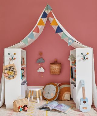 Kids bedroom painted a dark pink, two white storage shelves facing each other and supporting a canopy and bunting to create a cosy play area with a rug, natural storage baskets and colorful accessories and toys