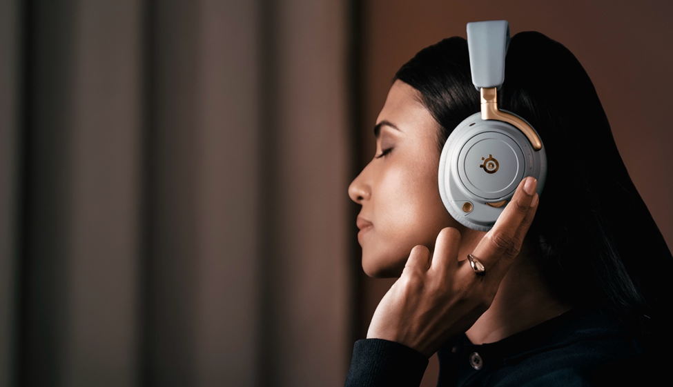 A woman listens to audio on the SteelSeries Arctis Nova Elite in the sage and gold colorway.