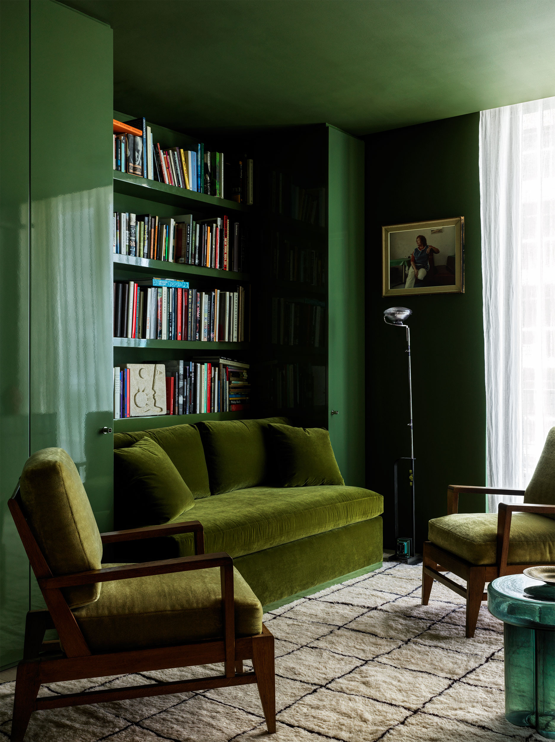 An all green living room drenched in color with green walls and ceiling, green velvet sofa and armchair, patterned rug, built-in bookshelf and sheer curtains