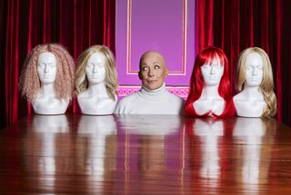 Sarah Beeny, who is bald and wearing a white polo neck in this picture, is positioned in the middle of four white busts wearing wigs of various lengths and colours