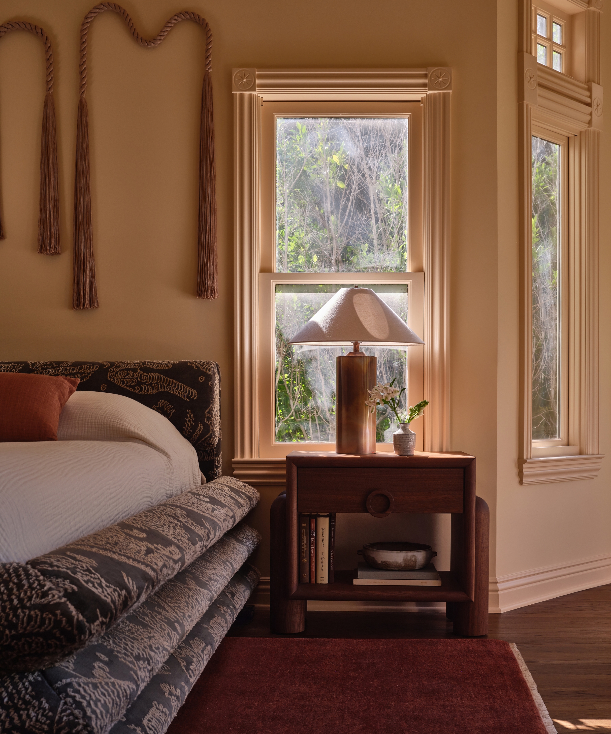 Warm-toned bedroom with cream walls and trim, a patterned bed, dark wood nightstand, sculptural lamp and rust-red rug.