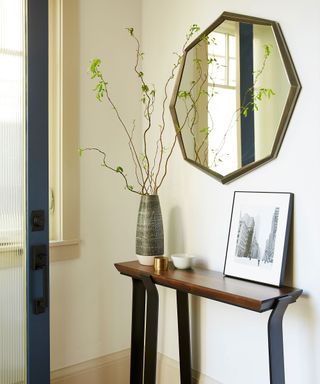 Cozy corner of an entryway with small console table, decorated with vase, framed picture and ornaments, octagonal mirror mounted above