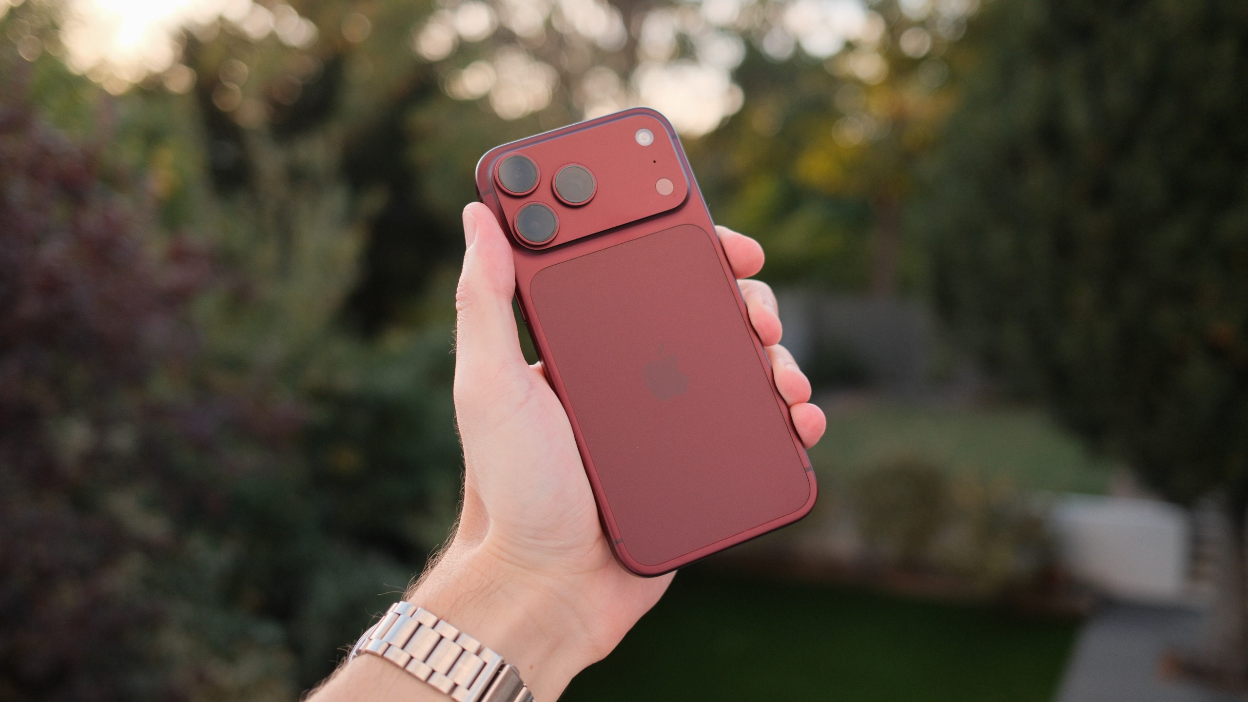 Burgundy iPhone 18 Pro Max held up in a garden, showing the rear design and triple-camera system.