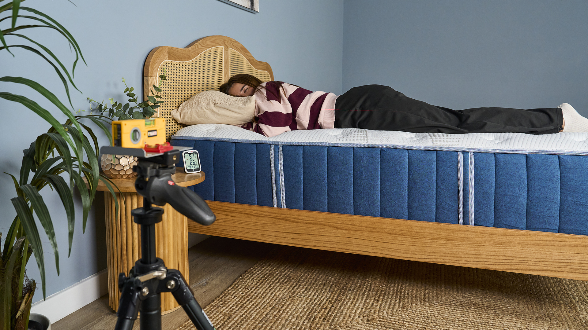 A wider shot of our mattress tester lying on their front on the navy and white Bear Elite Hybrid sitting on a wooden bed frame with a rattan headboard. They are wearing a striped sweater and black trousers. A laser on a tripod is set up in the foreground, facing the bed, projecting a red line on the mattress tester to show how well their body is aligned.