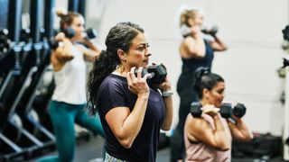 Woman holding dumbbell below her chin, about to squat with others in class, practising exercise for stress relief in the gym