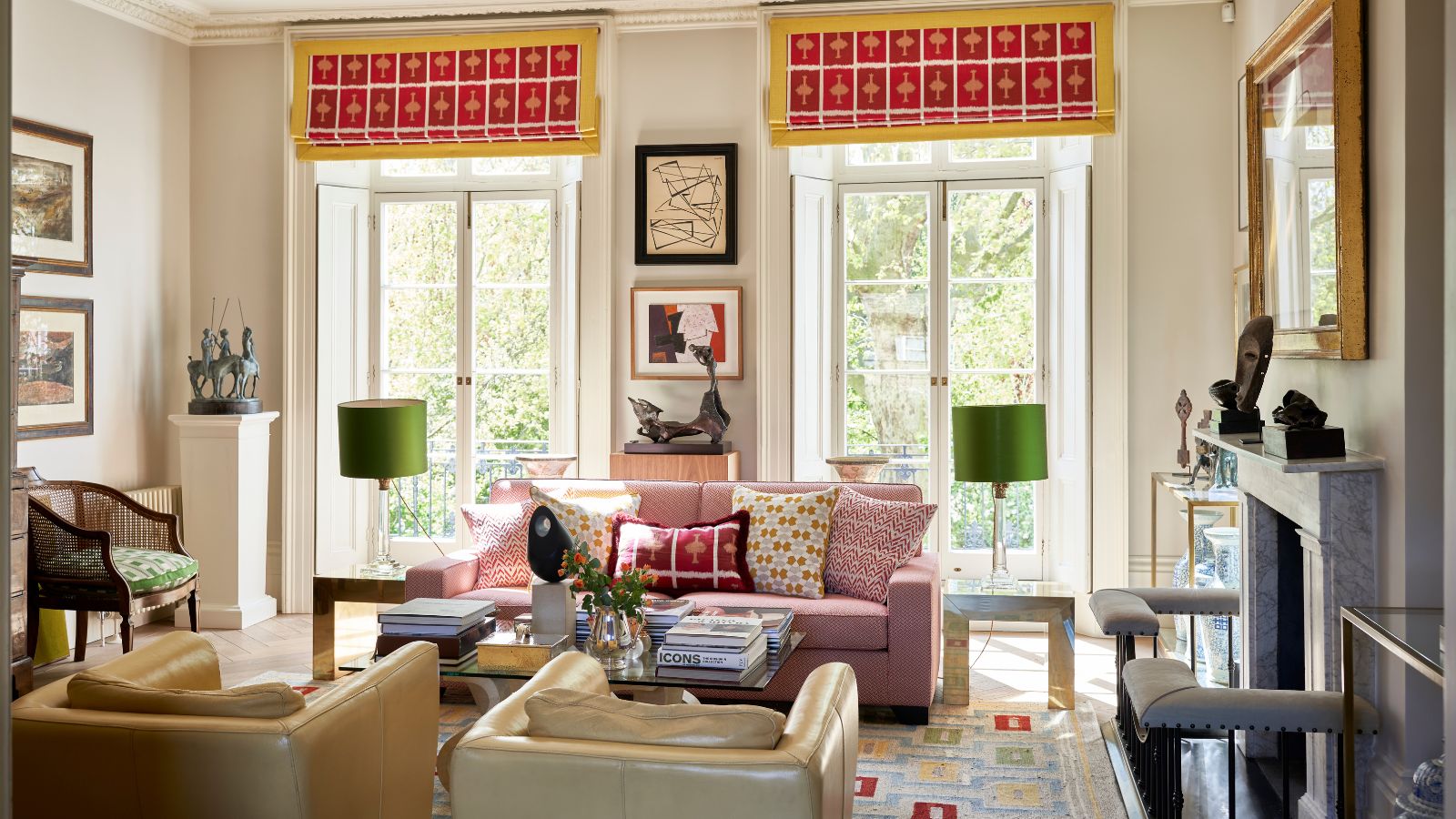 Bright, multi-colored living room with two large windows, a pin sofa, two leather armchairs, a coffee table with flowers and books, a patterned rug, and miscellaneous lights and framed pictures.