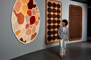 A young man dressed in a chequered blue and grey blazer, grey top and trousers stands in front of some colorful rugs hanging on a wall.