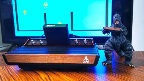 Atari 2600+ console sitting on woodgrain table with Berserk cart inserted and Godzilla toy holding tiny Atari console on right hand side