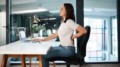 A woman sits at a desk holding her lower back and wincing.