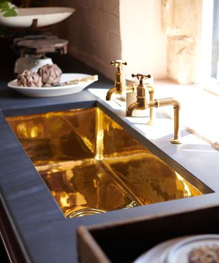 A striking kitchen sink crafted from polished brass, creating a reflective, golden surface. The sink is set into a dark, matte countertop and paired with traditional brass taps featuring porcelain cross handles.