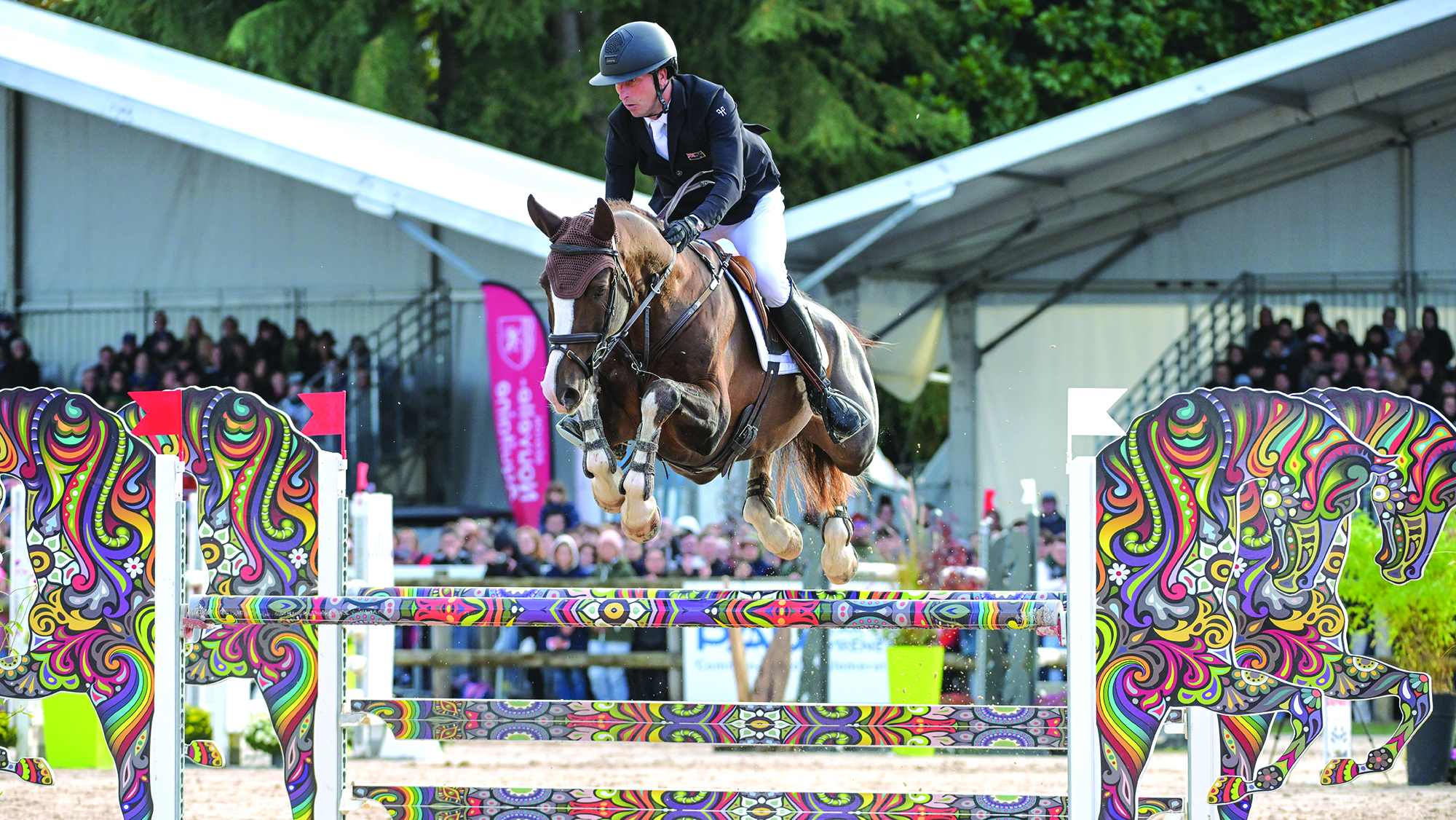A rider in a black coat rides a chestnut horse over a multi-coloured showjump at Pau Horse Trials 2025