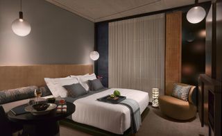 A room in the Nobu Hotel Shoreditch. The room is decorated in various shades of gray and brown- walls, carpet, and bedside tables are in lighter gray, while the armchair is in deep golden brown. The headboard is painted gold, with a rounded table next to the bed, which has food and vine on it. Floor-to-ceiling windows cover the far wall.