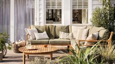 Green cushions on wooden patio furniture, in front of a white panelled home. 