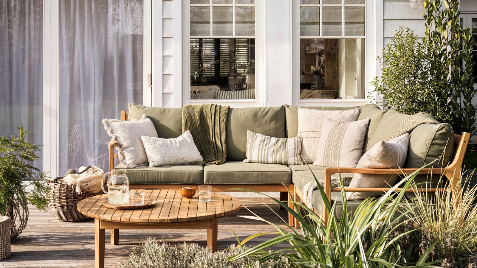 Green cushions on wooden patio furniture, in front of a white panelled home. 