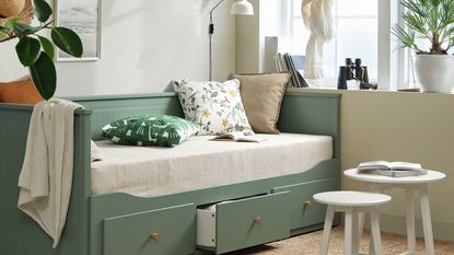 IKEA grey green HEMNES bed in a living room with floral cushions, large white windows, a pair of white round tables and a jute rug.
