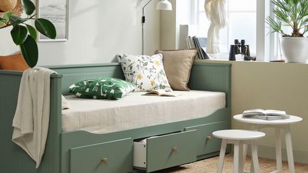 IKEA grey green HEMNES bed in a living room with floral cushions, large white windows, a pair of white round tables and a jute rug.