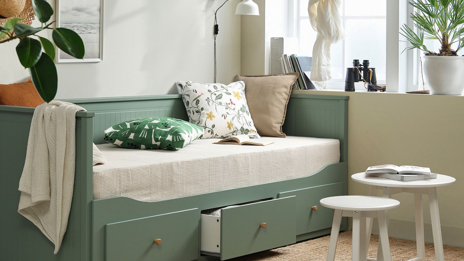 IKEA grey green HEMNES bed in a living room with floral cushions, large white windows, a pair of white round tables and a jute rug.