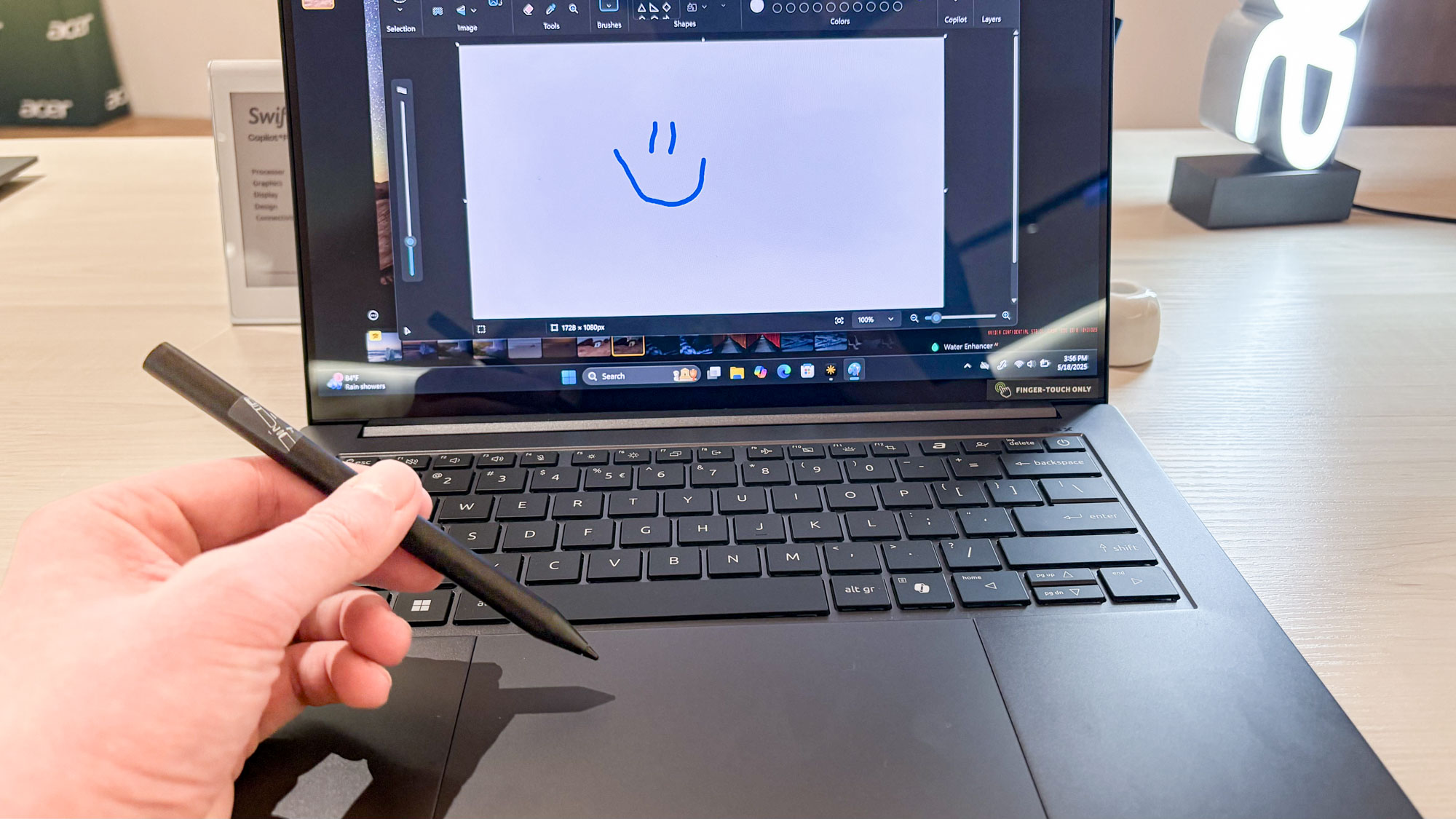 Hand holding Acer Stylus pen in front of Acer Swift X 14 with smiley face on display