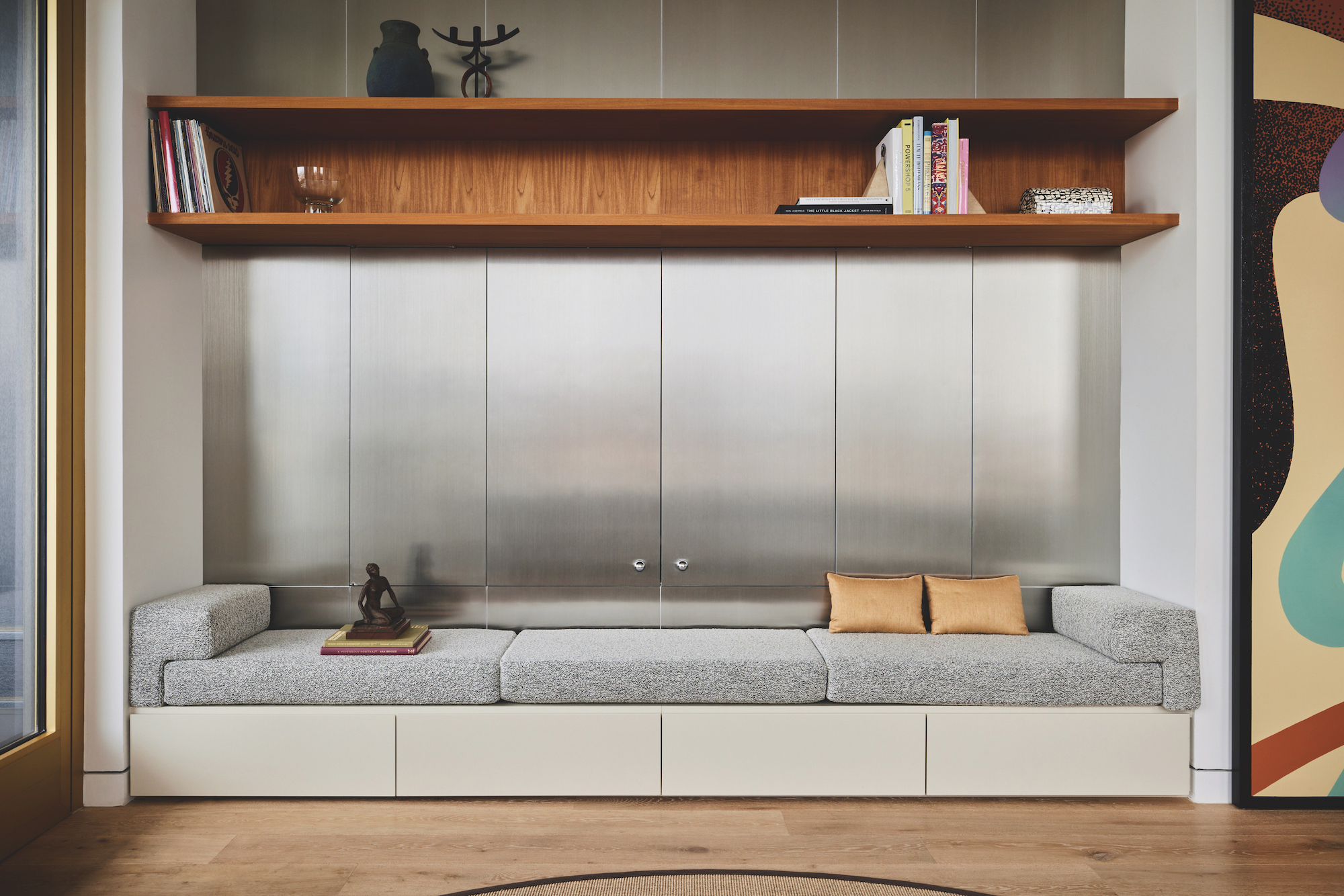A modern built in sofa with light grey upholstery and cream base, with metal clad walls behind and cedar coloured wood shelves above the sofa.